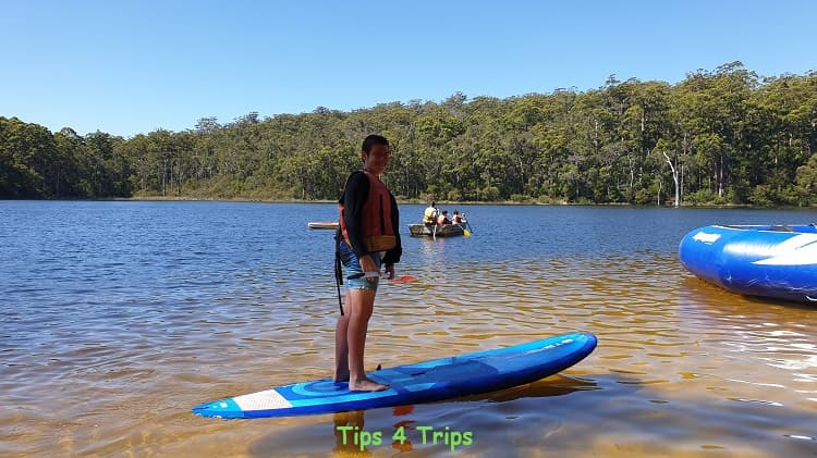 boy standing on stand up paddle board on lake at Karri Valley resort surrounded by trees